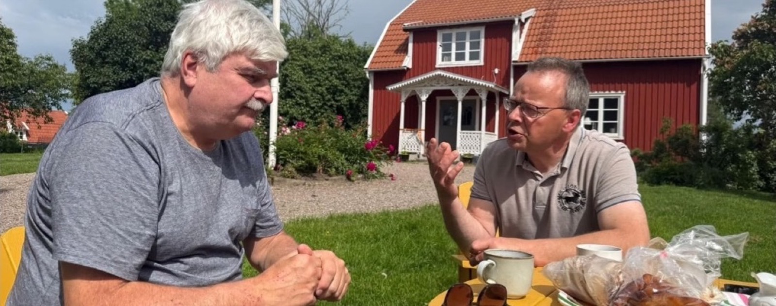 Erik i samtal över en kopp kaffe vid ett gult trädgårdsbord framför en röd gård